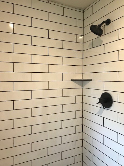 Shower wall in long white tile with black grout, corner shelf and black fixtures